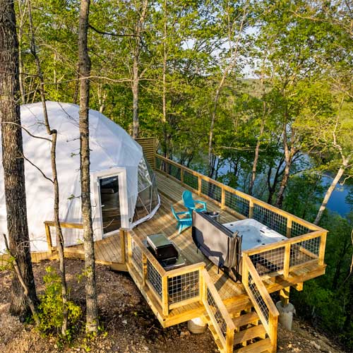 Aerial view of a glamping dome and tiered wooden deck featuring a hot tub, grill, and lake view nestled among the trees.
