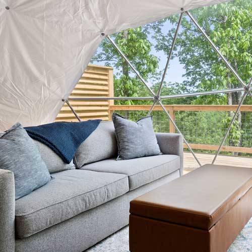 Cozy interior of a glamping dome with a plush gray sofa, leather ottoman, and panoramic forest views through a geodesic window.