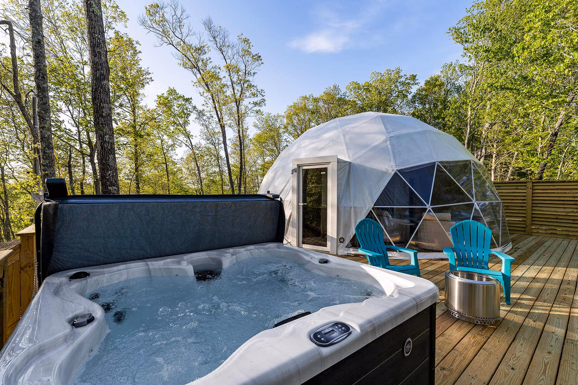 Outdoor view of a glamping dome near Center Hill Lake featuring a bubbling hot tub, two turquoise Adirondack chairs, and a private deck surrounded by trees—perfect for a luxurious nature escape.