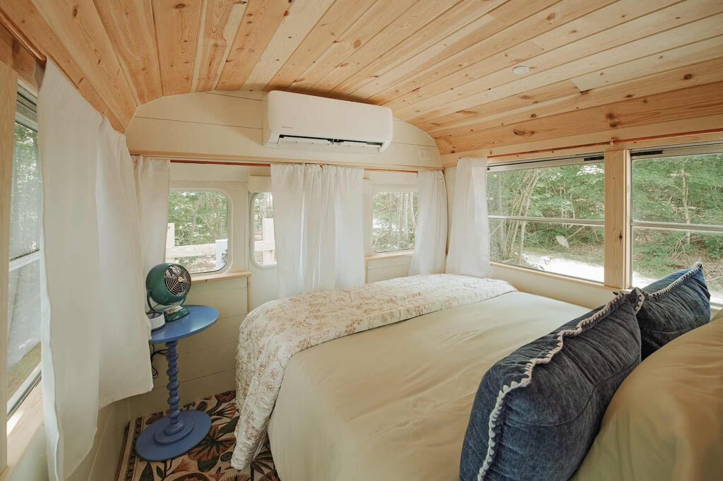Bedroom inside a skoolie rental in Tennessee featuring a plush bed, wood-paneled ceiling, and wraparound windows with forest views.