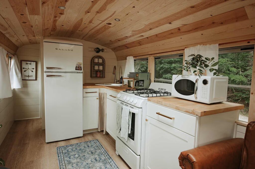 Kitchen inside a skoolie rental in Tennessee with full-size fridge, gas stove, microwave, and natural wood finishes.