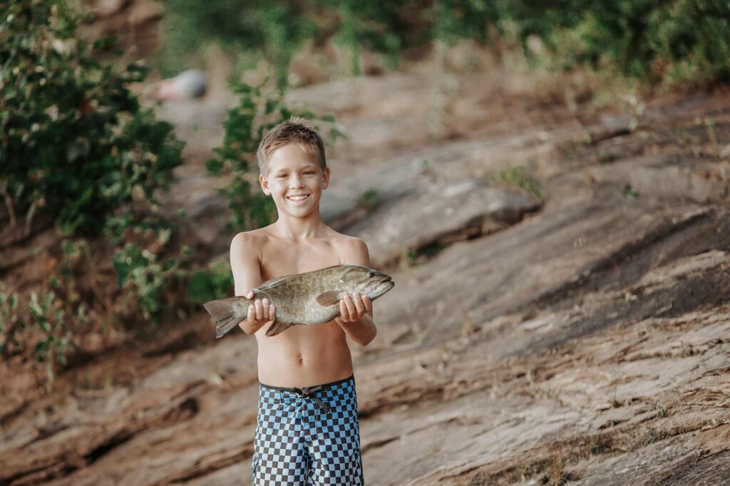 Smiling boy proudly holding a freshly caught smallmouth bass on the rocky shoreline of Center Hill Lake—perfect for beginner anglers.
